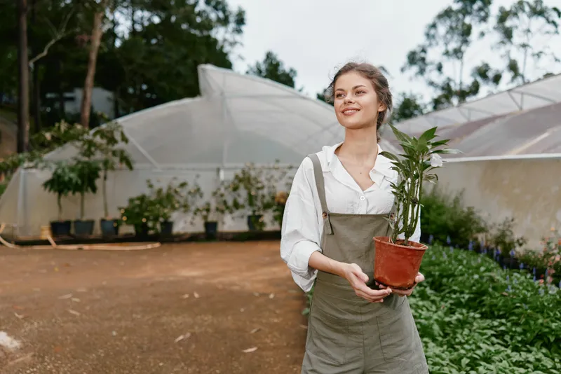 Young woman smiling while holding a potted plant in a greenhouse, showcasing a connection to nature and gardening, perfect for promoting eco friendly practices
