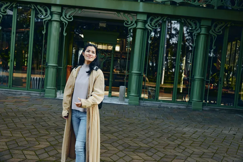 Woman wearing a long beige coat stands outdoors in front of green glass doors, casual denim and sweater visible, framed by arches and a tiled courtyard beside architectural detail
