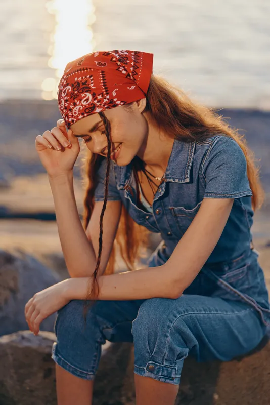 Lifestyle woman in modern boho-western denim street style wearing a red bandana, enjoying a warm sunset with film color tones near water background.