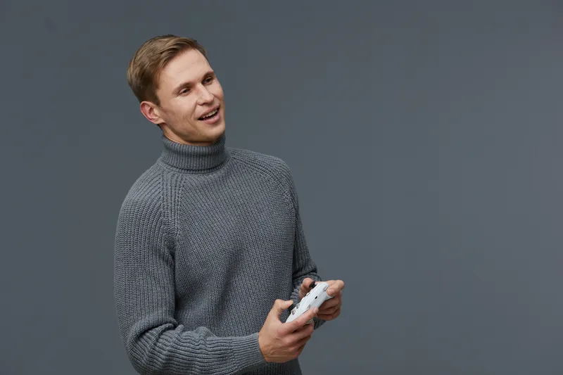 Smiling young man with blond hair wearing a cozy gray knit sweater holding a mobile phone, standing against a plain gray background. Casual fashion and technology concept.
