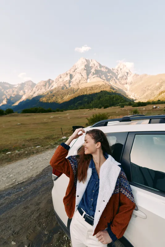 Woman by car with mountains landscape, travel road and nature portrait during golden hour. Cozy jacket, smiling and relaxed, outdoor adventure scene with scenic valley and alpine peak background.