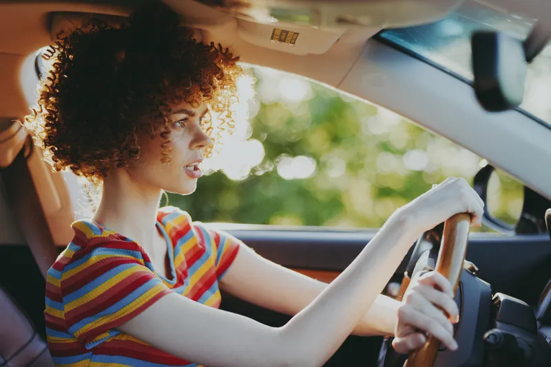 Vibrant woman driver in a colorful striped shirt sits in a car outdoors, sunlight framing her profile, a joyful, relaxed moment captured with retro film color and warm natural tones mood light