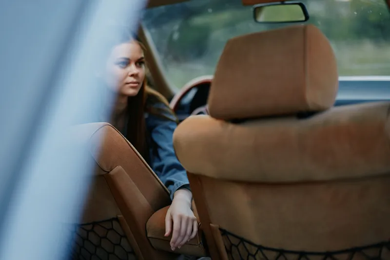 Young woman sitting in a car, looking thoughtfully out the window, with a soft brown interior and natural green background. Reflects emotions of contemplation and serenity.
