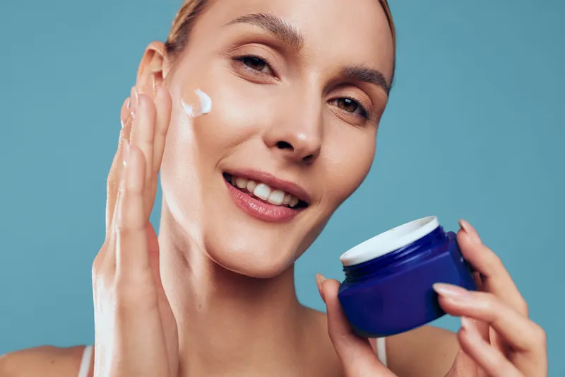 A smiling woman applies moisturizing cream to her cheek holding a blue jar. Close-up of skincare routine and beauty concept on a blue background with natural makeup.