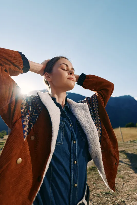 Woman, portrait and outdoors with sunlight and jacket near mountains, enjoying nature and travel. Female model in denim shirt with closed eyes, relaxed pose in countryside golden hour.