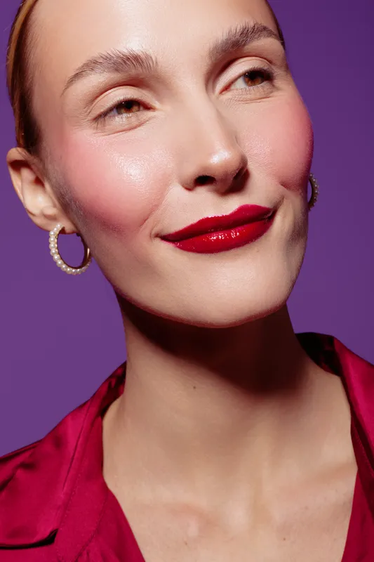 Close-up portrait of a confident woman with red lipstick and pearl earrings wearing a red blouse against a purple background. Concept of beauty, fashion, and elegance.