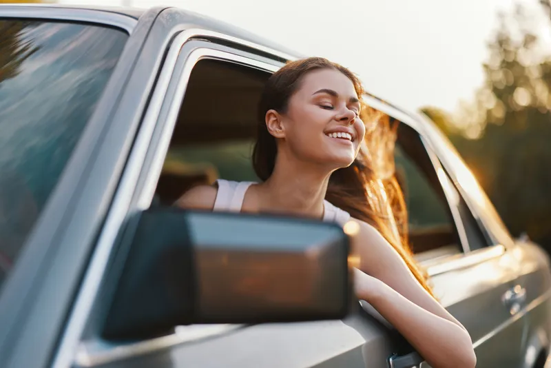 Happy young woman enjoying a sunny day, leaning out of a car window, with sunlit hair, showcasing freedom and joy during a road trip.