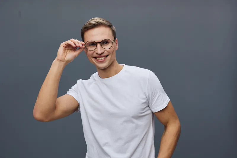 Smiling young man with glasses in casual white t-shirt adjusting his spectacles and looking at camera isolated on plain gray background studio portrait. People casual lifestyle concept