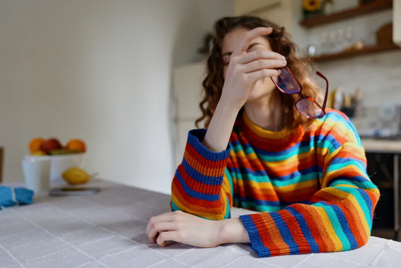 A young woman in a colorful rainbow striped sweater sits at a kitchen table, resting her head on one hand and holding glasses, conveying fatigue and reflection.