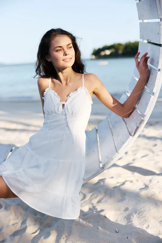 A graceful woman in a white sundress stands by a curved white barrier on a sunlit beach, exuding calm charm, soft shadows, and relaxed confidence.