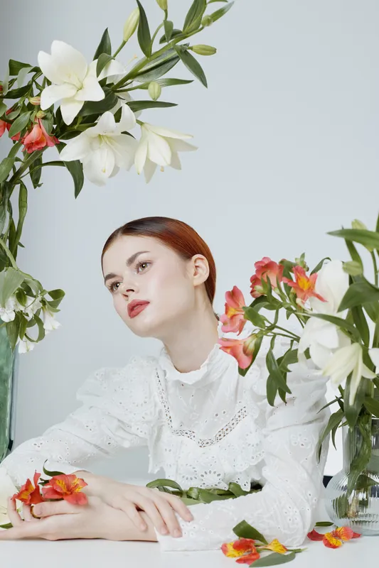 Elegant young woman in a white lace blouse surrounded by fresh flowers, expressing serenity and beauty in a soft, pastel toned setting