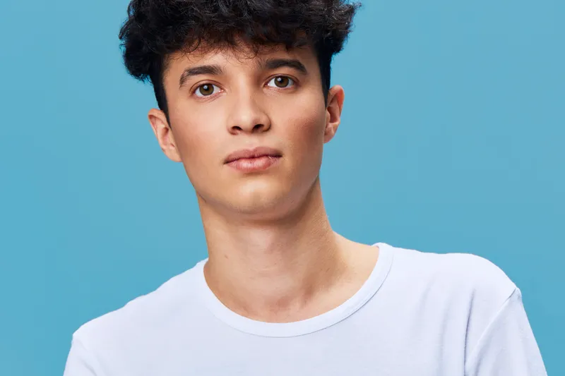young man portrait with curly hair wearing casual white t-shirt standing against blue background, looking calm and confident with natural expression