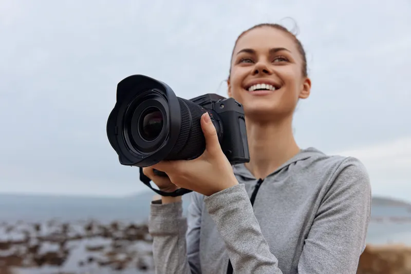 Joyful young woman holding a camera by the beach, capturing photography memories, wearing casual sporty clothing with a grey palette, ideal for lifestyle concepts