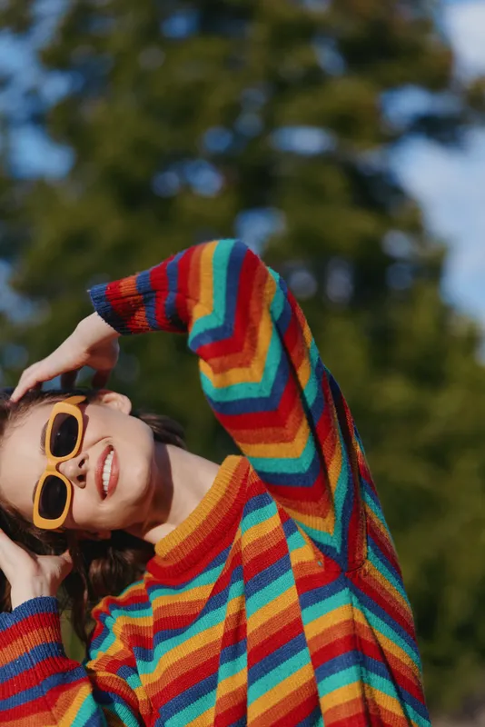 Woman smiling in striped sweater and sunglasses outdoor. Young adult model in casual outfit leaning back with joyful expression, colorful fashion portrait for lifestyle and travel use.