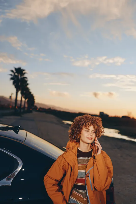 Woman lifestyle at sunset, standing near car on coastal road, wearing orange jacket and striped top, curly hair blowing, relaxed expression, cinematic evening light by beach.
