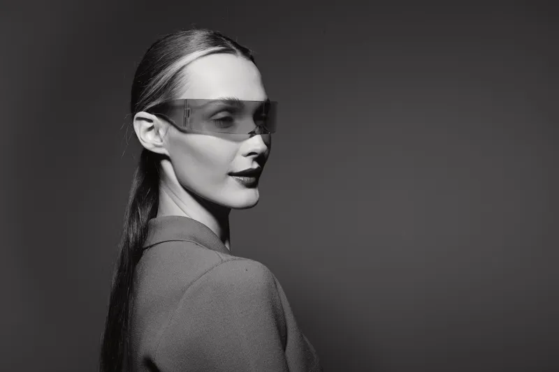 Elegant caucasian woman with light skin wearing futuristic glasses and textured jacket poses in studio with monochrome background.
