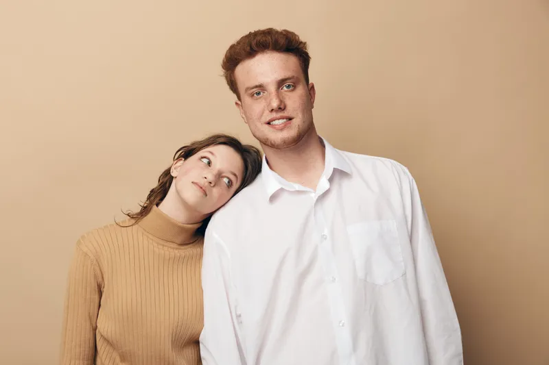 Young couple with woman leaning on man shoulder, both smiling gently. Casual clothing and neutral background create warm, intimate, and relaxed mood. Concept of love, friendship, and connection.