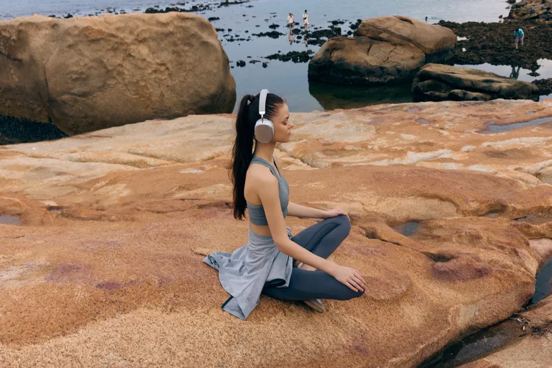 Young woman in yoga pose with headphones on rocks by the sea, promoting mindfulness and relaxation amid a serene coastal environment