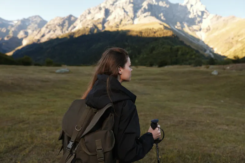 A lone hiker with a backpack gazes toward rugged mountains under a clear sky, capturing a moment of adventure and exploration in a vast alpine landscape.