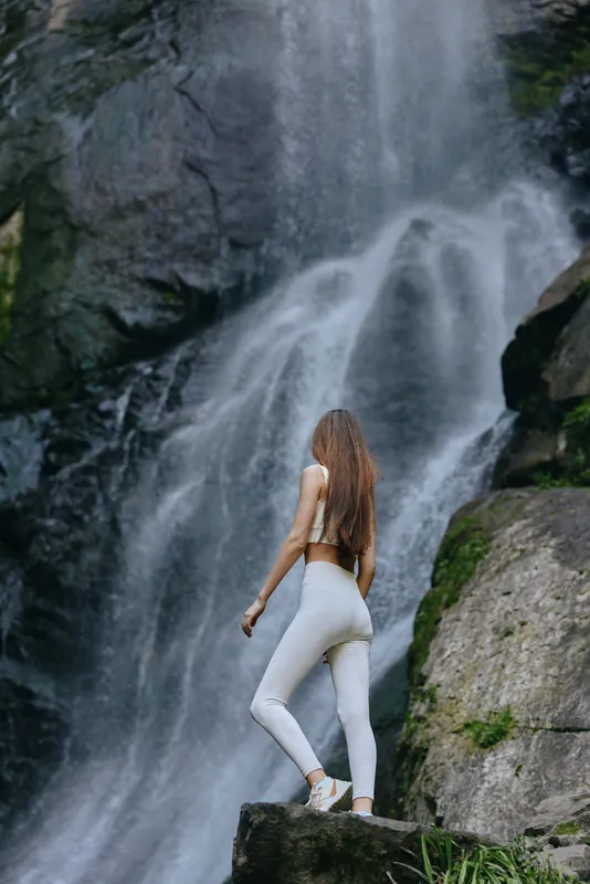 woman standing near waterfall in activewear, long hair, natural outdoor setting with rocks and flowing water, fitness and adventure concept