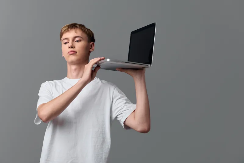 Laptop held aloft by casual youth against a neutral backdrop, emphasizing technology and presentation as a modern display of digital skill and portable device use