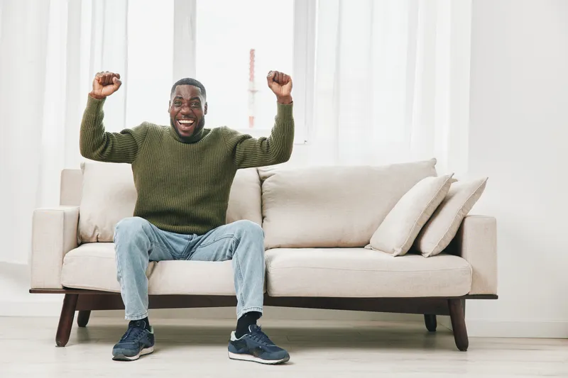 Happy African American man celebrating on a couch, wearing a green sweater and blue jeans, exuding joy and excitement in a bright living room