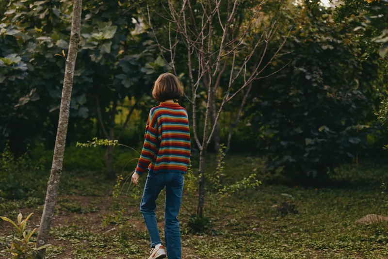 Preteen boy, 10 year old child walking through forest nature in a colorful striped sweater and jeans, casual outfit with sneakers, thoughtful mood, outdoor lifestyle travel and exploration.