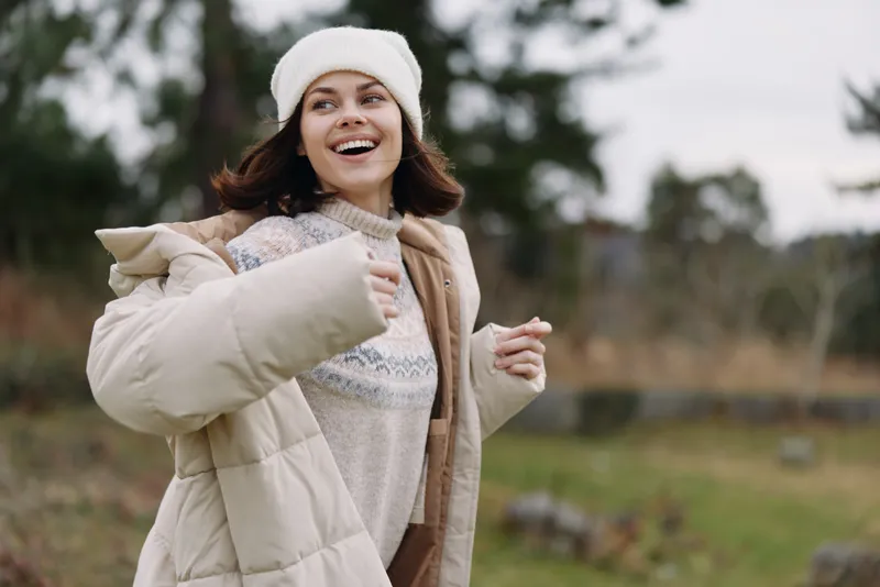 Woman outdoors wearing a winter coat, smiling, with scarf and beanie in a warm outdoor setting, showcasing cozy fashion and carefree mood in a natural landscape.
