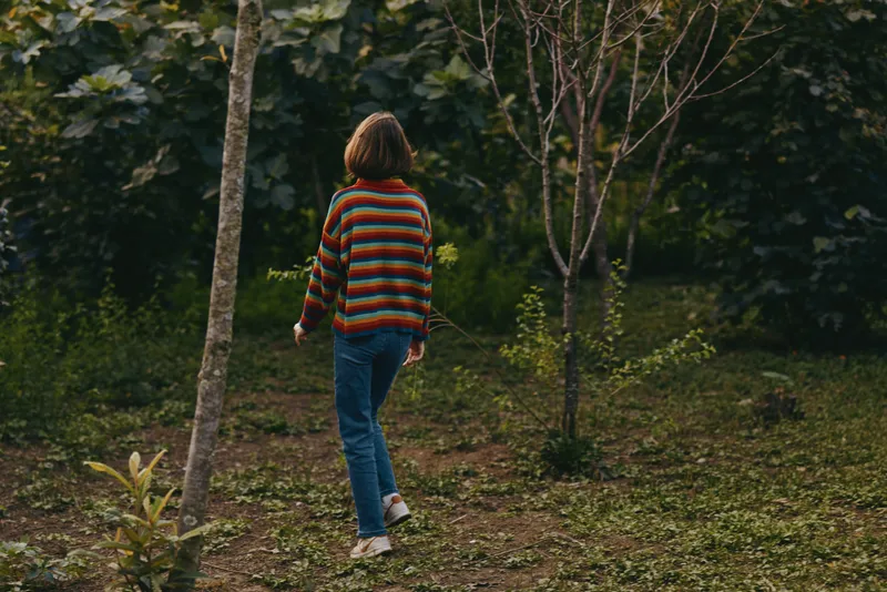 Young woman in a striped sweater walking away through a forest garden, casual outfit with jeans and sneakers and a short bob haircut, contemplative mood for lifestyle or travel scene.