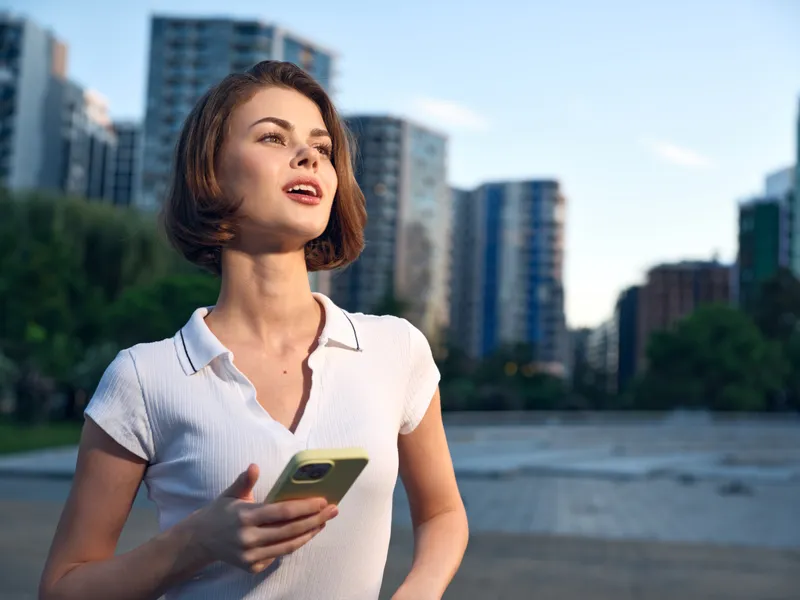 A cheerful businesswoman holds a smartphone in a modern outdoor setting with tall buildings in the background. She smiles and appears engaged, creating a positive urban atmosphere.