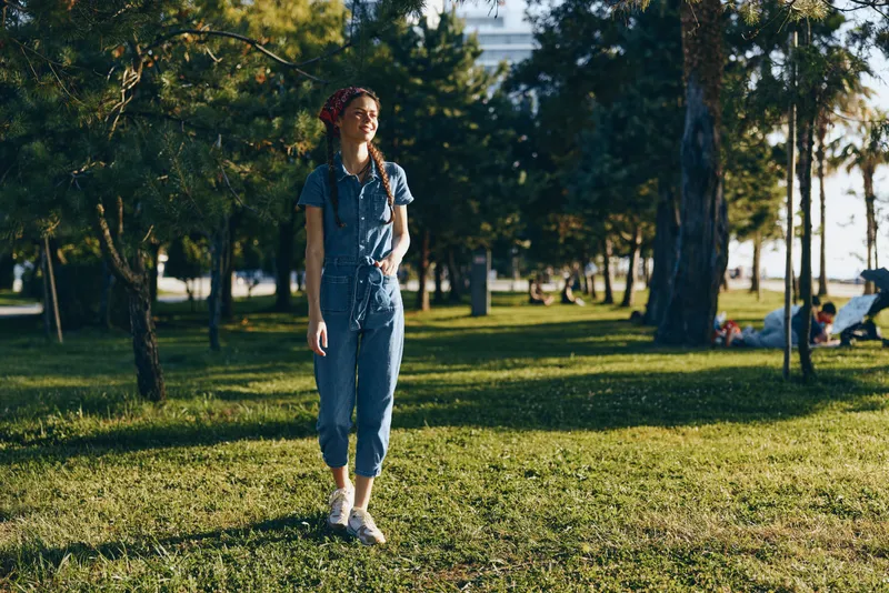 young woman walking in park wearing denim jumpsuit with braided hair enjoying sunny day and green grass in background