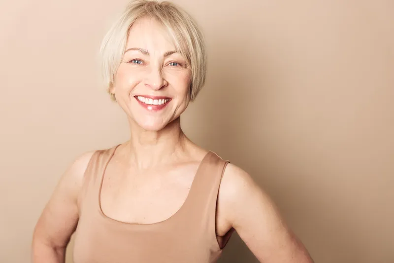 Smiling mature woman with short blonde hair wearing beige sleeveless top stands confidently against neutral background. Concept of happiness, aging, beauty, confidence, and wellness.