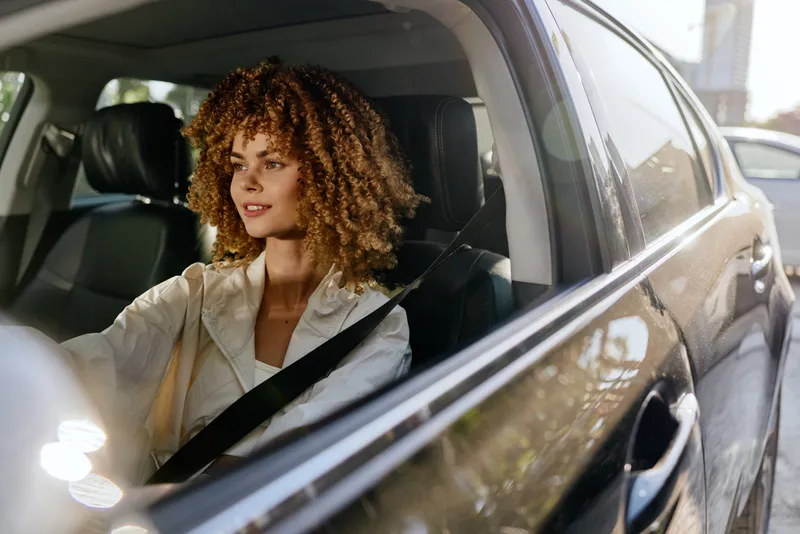 Smiling woman with curly hair driving a car, wearing a beige jacket and seatbelt, enjoying sunny weather, urban background visible through windows.