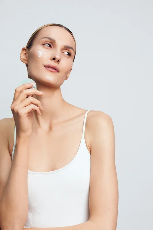 Young woman with light skin applies face cream on her cheek while holding a cosmetic jar against a plain light background in a calm skincare routine.