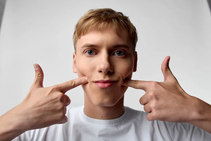 Smiling young man pulls at the corners of his mouth with both fingers, creating a playful grin in a clean studio setting, wearing a white tee.