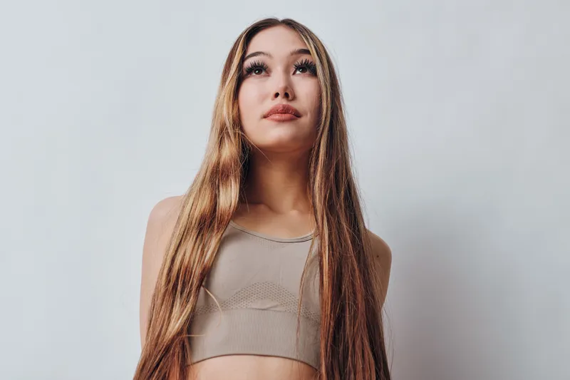 Young woman with long straight hair posing in beige crop top looking thoughtfully upward against light background for beauty and fashion concept.