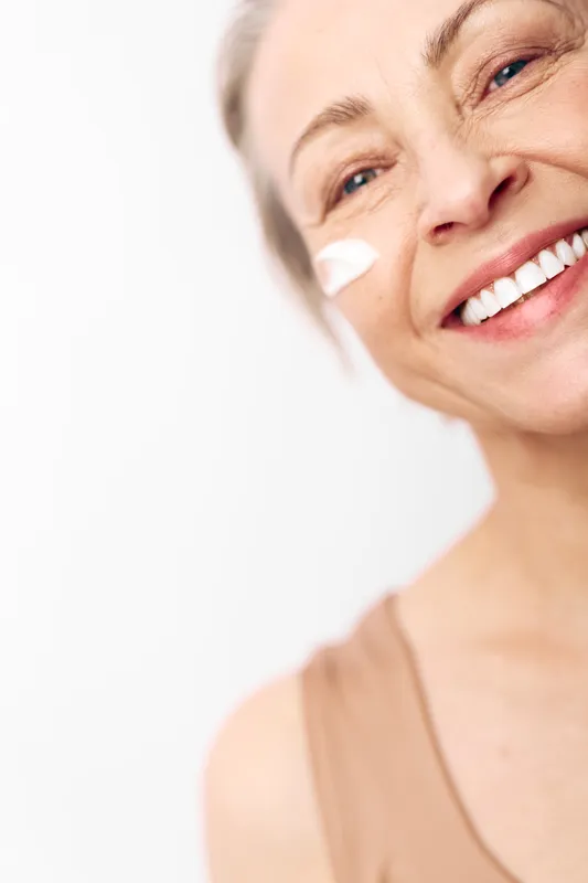 Smiling mature woman applies white face cream on cheek, showing healthy skin and joyful expression. Close-up portrait on bright background with natural light for skincare concept.