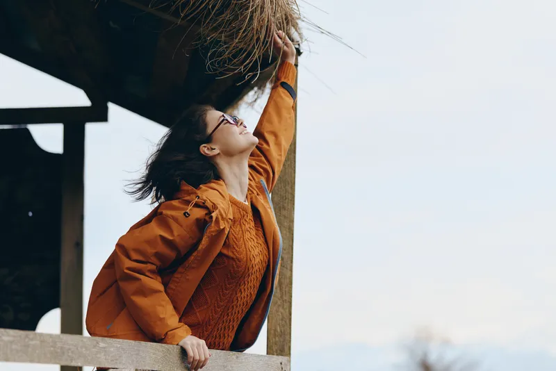Woman outdoor lifestyle jacket hair wind railing smile portrait, leaning on wooden railing in warm daylight, hair blown by breeze, blurred sky background, casual fashion mood
