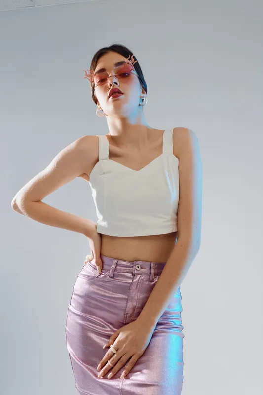 Stylish woman in a white crop top and shiny pink skirt poses confidently against a plain background. Concept of beauty, fashion, and modern style with elegant accessories.