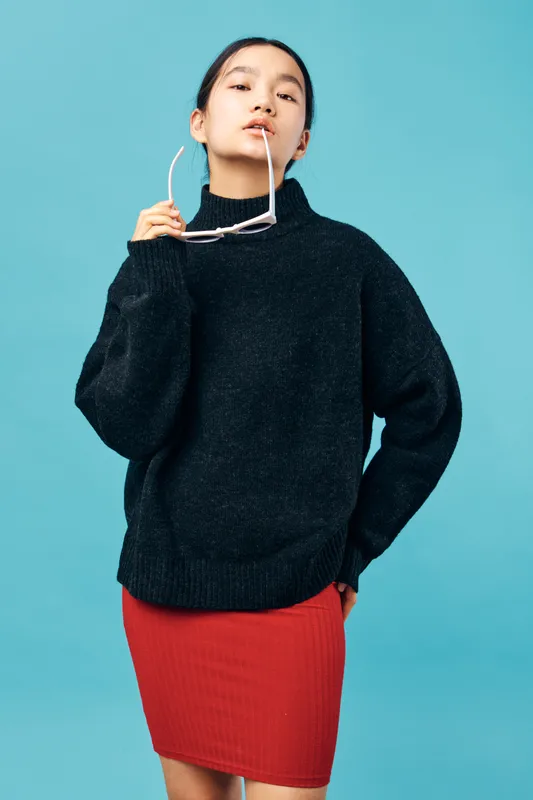 Young woman posing with sunglasses in hand wearing black sweater and red skirt against blue background confident and stylish with casual fashion look and modern attitude.
