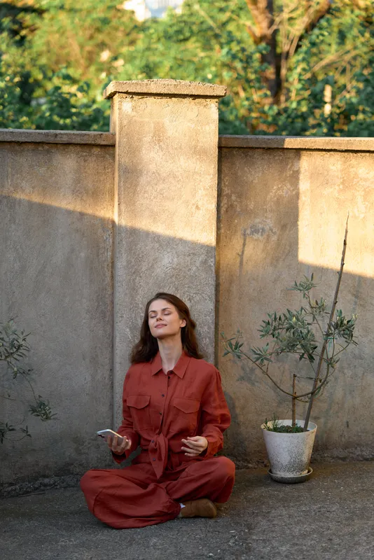 A woman in a rust red dress sits cross legged, meditating in a sunlit courtyard with a tall wall, potted plant, and warm light. Concept: mindfulness, calm, lifestyle balance