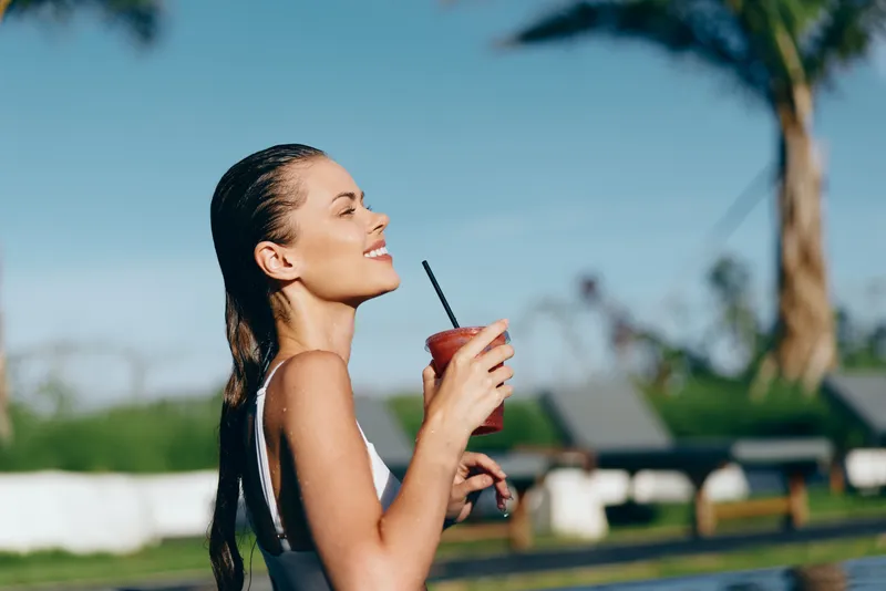 A young woman in a swimsuit enjoys a refreshing drink outdoors near a pool on a sunny day, embodying a relaxed lifestyle and summer leisure concept.