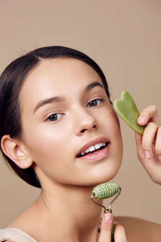 Young woman demonstrating facial massage techniques with jade tools on a beige background, emphasizing wellness and beauty in a serene atmosphere
