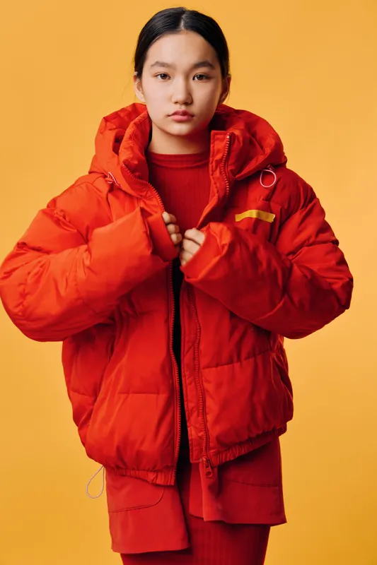 Woman wearing bright orange puffer jacket and matching red outfit standing against yellow background. Casual winter fashion, portrait, cool style, confident look with hands adjusting zipper.
