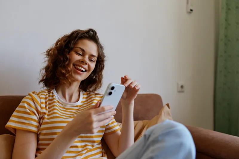 A young woman relaxes on a sofa in a striped yellow t shirt, smiling at her phone for a casual home lifestyle moment.