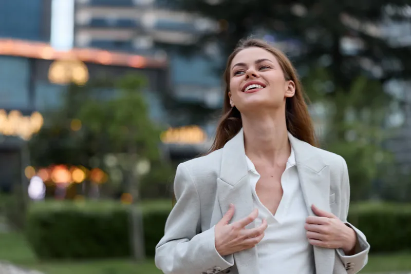 smiling woman in light blazer enjoying outdoor urban environment with natural green background and soft evening light