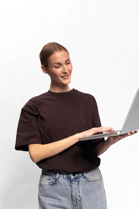 Young woman in casual clothes smiling while holding and using laptop. White background with soft lighting creates clean and simple technology concept image.