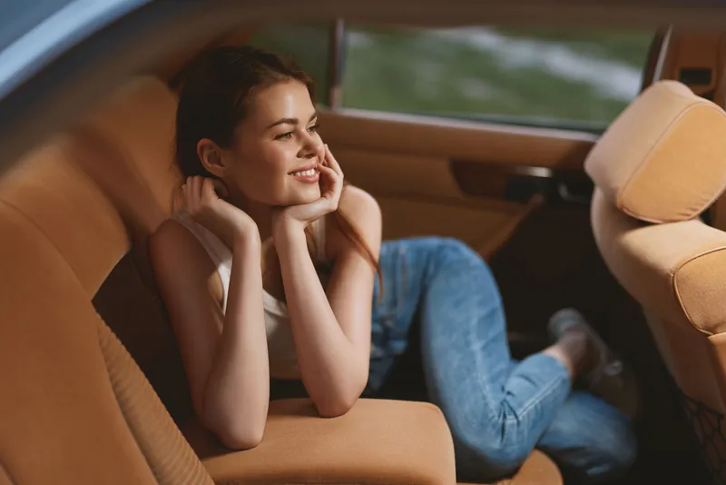 Young woman relaxing in a car, smiling brightly, wearing a casual outfit, upholstered in soft tan fabric, evoking a sense of joy and freedom.