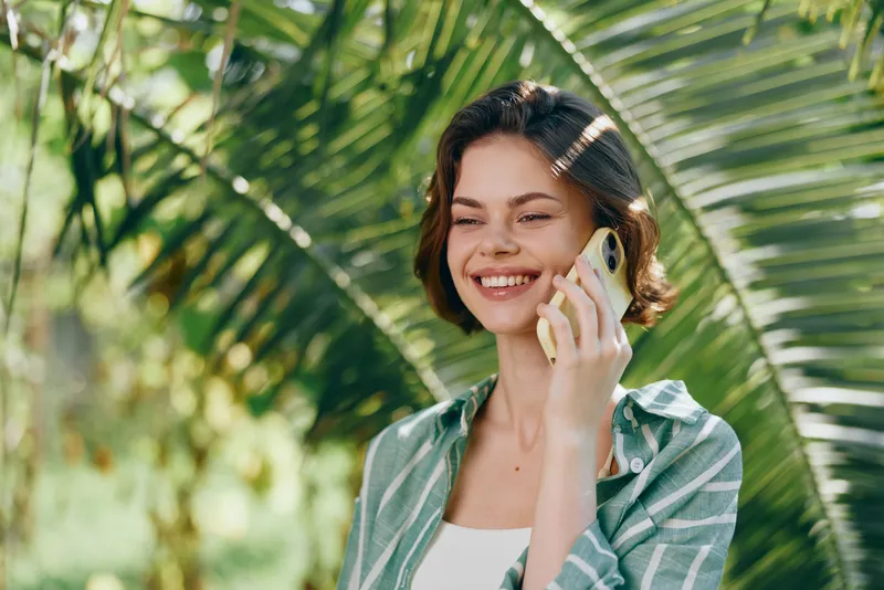 Smiling woman talking on a mobile phone outdoors among tropical greenery, warm sunlight highlighting her cheerful expression.
