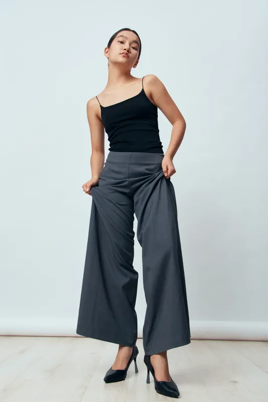 Young stylish woman posing in loose gray pants and black tank top with high heels in white studio. Female model confident and elegant with casual fashion outfit and modern style look.